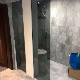 Szklana ścianka kabiny prysznicowej zamontowana w nowoczesnej łazience z szarą mozaiką na ścianach i koszem wiklinowym wewnątrz kabiny.