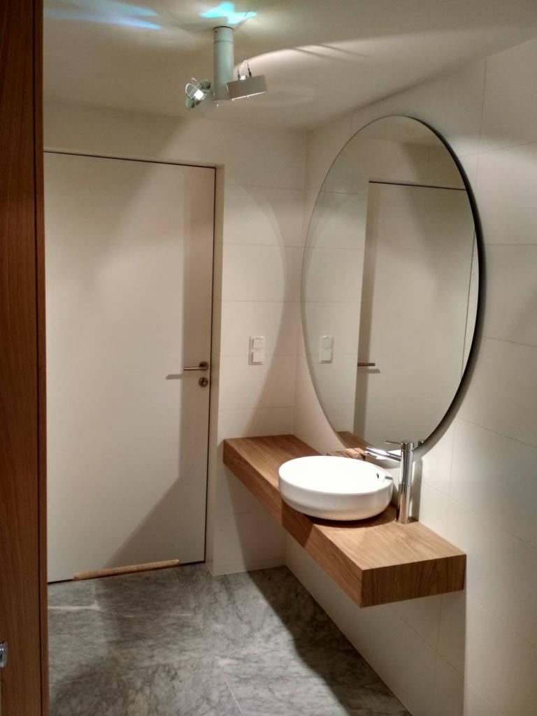 Nowoczesna łazienka z owalnym lustrem, białą umywalką nablatową na drewnianym blacie, szare płytki podłogowe i sufitowe oświetlenie punktowe.