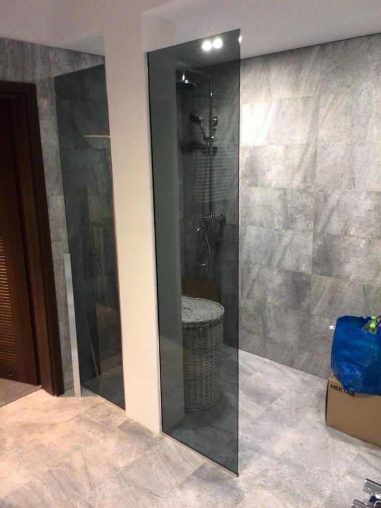 Szklana ścianka kabiny prysznicowej zamontowana w nowoczesnej łazience z szarą mozaiką na ścianach i koszem wiklinowym wewnątrz kabiny.