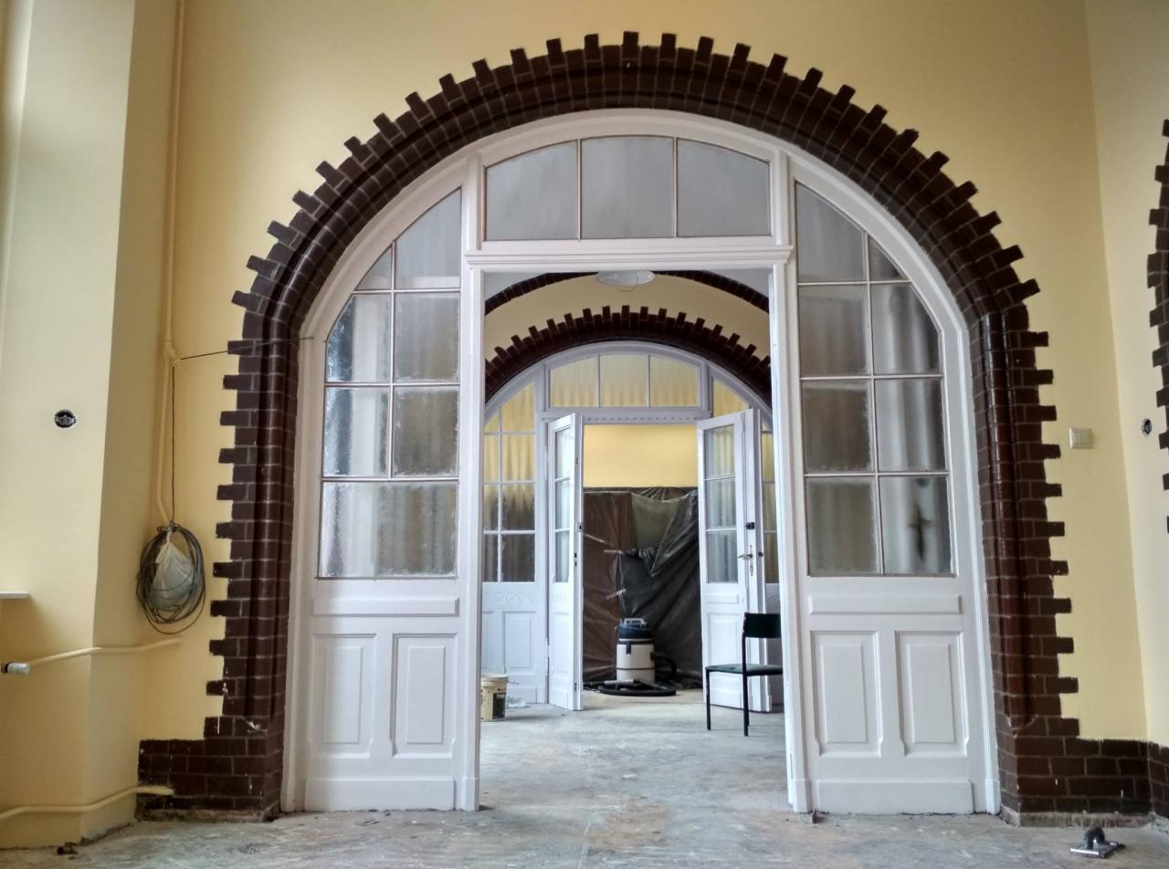Wysokie, przeszklone, białe drzwi łukowe z ceglaną obudową w trakcie prac wykończeniowych, widoczne narzędzia i sprzęt sprzątający.