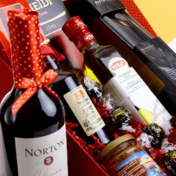 Zestaw prezentowy z winem w butelce z czerwoną kokardą w białe kropki, oliwą z rozmarynem, syropem malinowym, czekoladkami Lindor i włoskim sosem w słoiku, ułożony na czerwonej bibule.