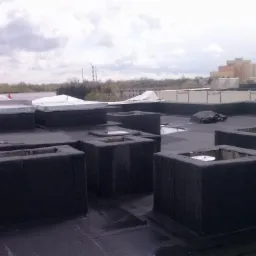 Pracownik w kasku na płaskim dachu pokrytym czarnym materiałem bitumicznym z licznymi kwadratowymi elementami wentylacyjnymi; w tle budynki i pochmurne niebo.