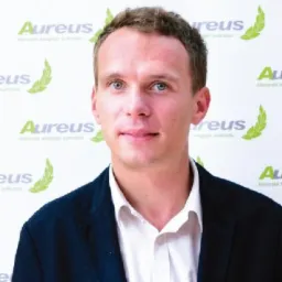 Portret mężczyzny w ciemnej marynarce i białej koszuli na tle z logo Aureus, z delikatnym uśmiechem i jasnymi oczami.