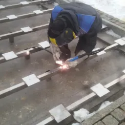 Pracownik w kombinezonie i masce spawalniczej łączy metalowe profile kwadratowe za pomocą spawarki na zewnątrz, widoczne białe płytki mocujące i śnieg na obrzeżach.