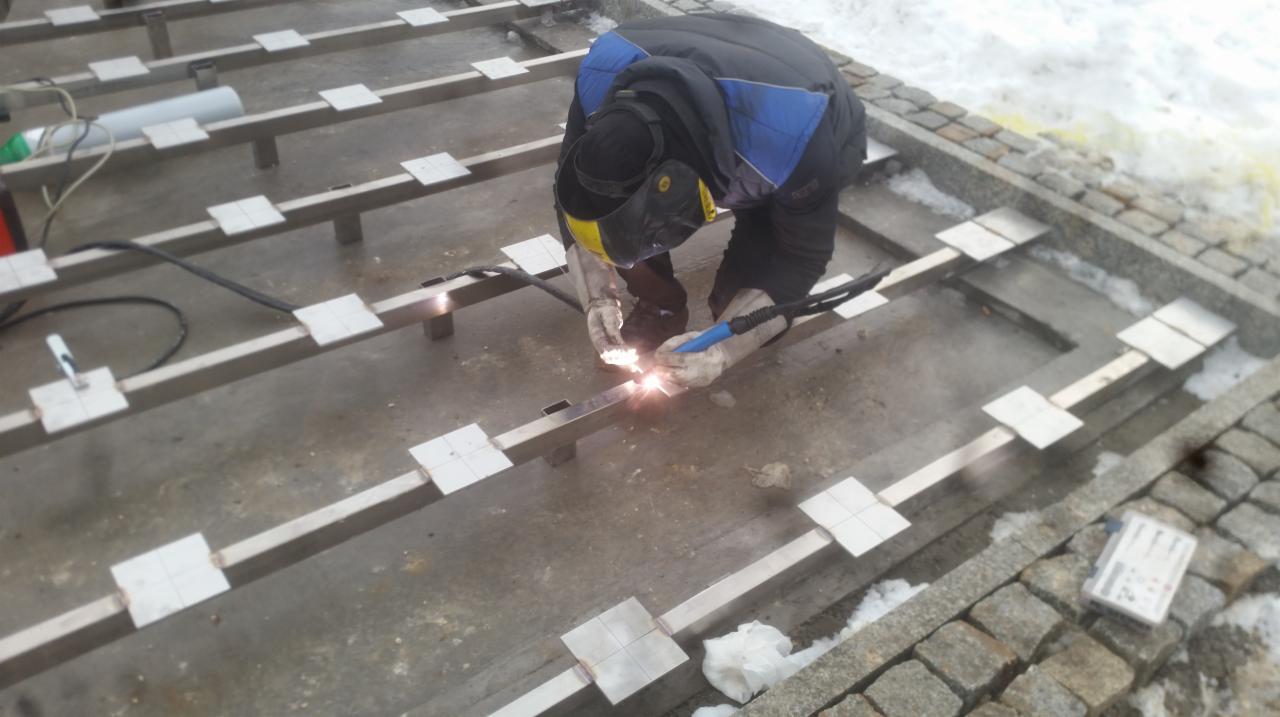 Pracownik w kombinezonie i masce spawalniczej łączy metalowe profile kwadratowe za pomocą spawarki na zewnątrz, widoczne białe płytki mocujące i śnieg na obrzeżach.