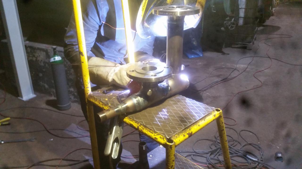 Spawanie rurociągu z kołnierzami na stanowisku roboczym, widoczny spawacz w masce i rękawicach ochronnych, butla z gazem w tle.