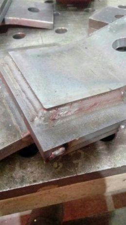 Metalowe elementy konstrukcyjne z widocznymi spawami narożnymi, leżące na perforowanym stole spawalniczym, z widocznymi odpryskami spawalniczymi.