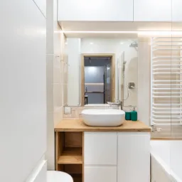 Nowoczesna łazienka z umywalką nablatową na drewnianym blacie, białe szafki z frontami bez uchwytów, widoczne WC i fragment wanny, w lustrze odbicie kuchni.