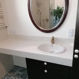 Łazienka z owalnym lustrem w drewnianej ramie, blatem z jasnego konglomeratu, umywalką i czarną szafką z białymi gałkami; w lustrze odbija się kabina prysznicowa z złotą armaturą i roślina doniczkowa.