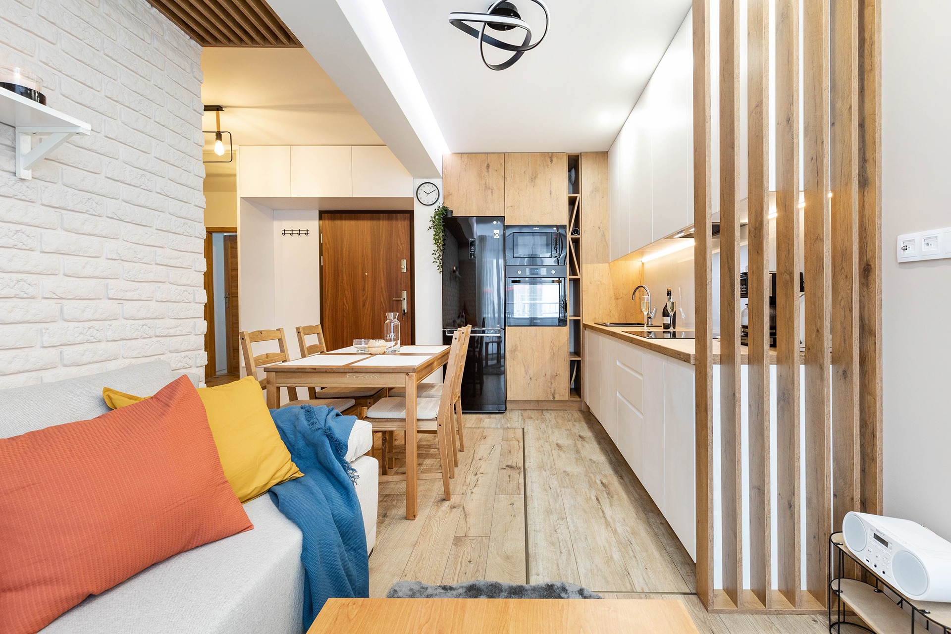 Jasne, nowoczesne wnętrze: kuchnia z drewnianymi akcentami, białe szafki, czarna lodówka, stół jadalny z krzesłami, sofa z kolorowymi poduszkami i drewniana ażurowa ścianka działowa.