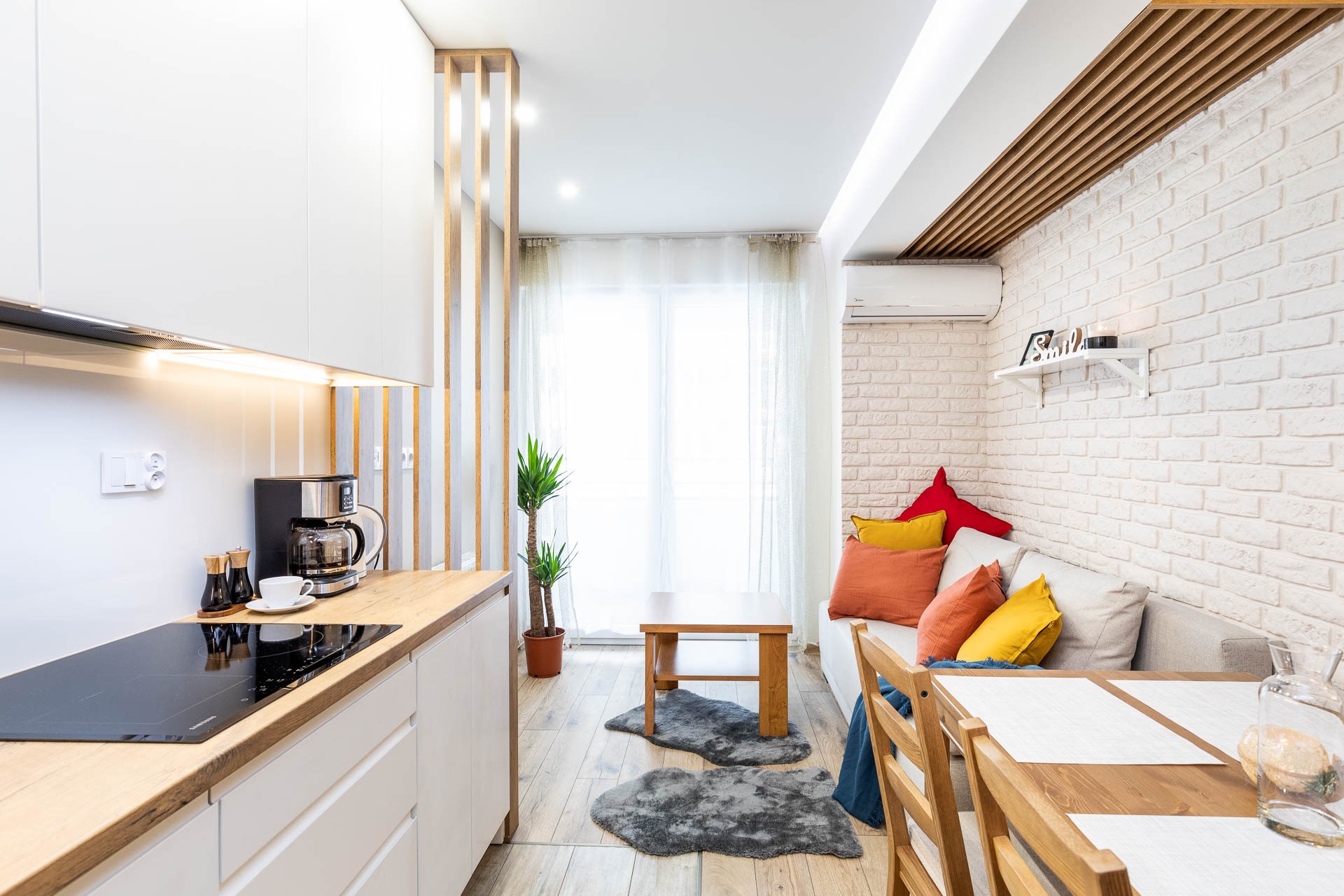 Jasne wnętrze mieszkania z elementami drewna: białe szafki kuchenne z drewnianym blatem, płyta indukcyjna, ekspres do kawy, salon z ceglaną ścianą, drewniany stół i krzesła, sofa z kolorowymi...