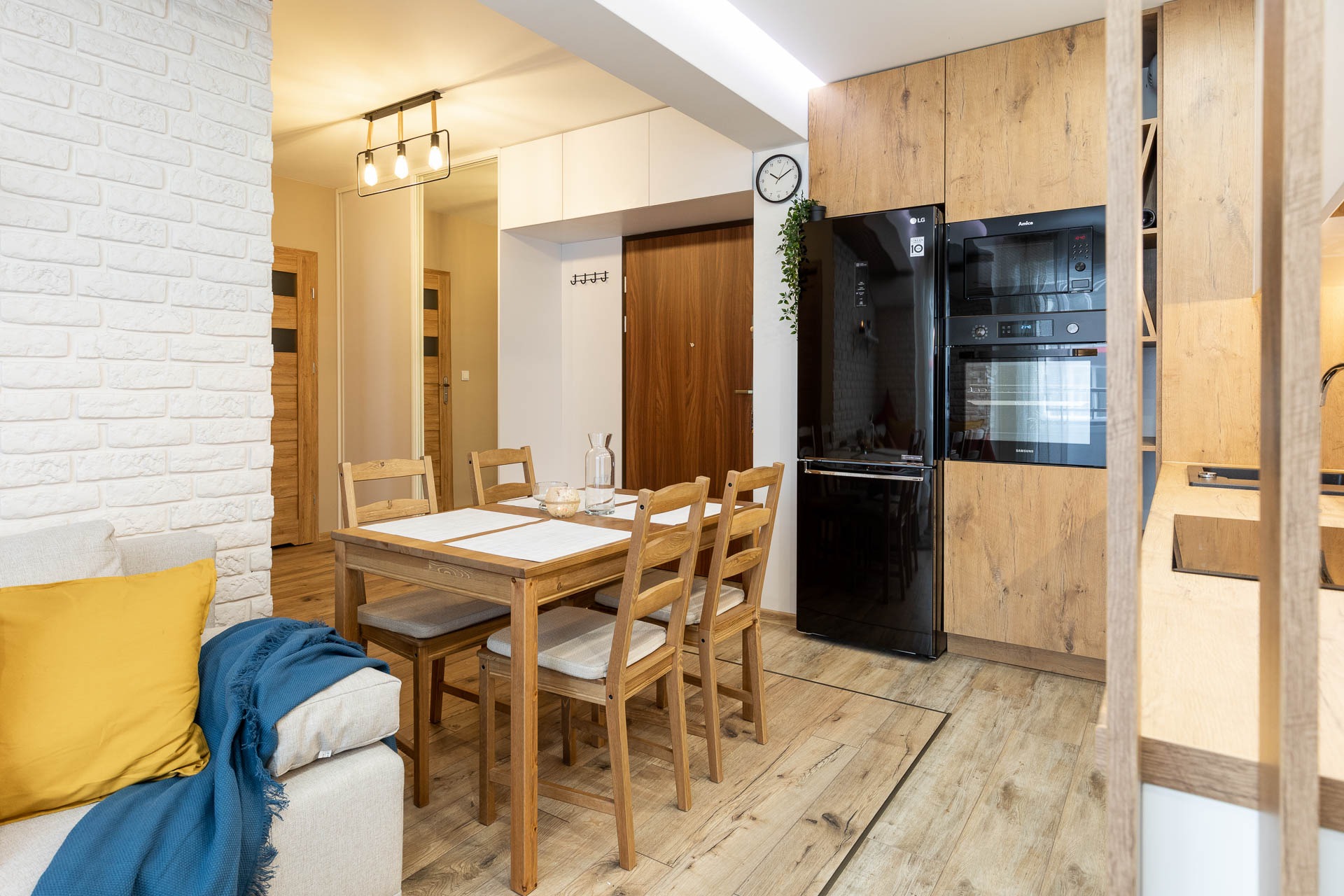 Jasna kuchnia z drewnianymi meblami, czarną lodówką i piekarnikiem w zabudowie, widok na stół z krzesłami i ścianę z białej cegły