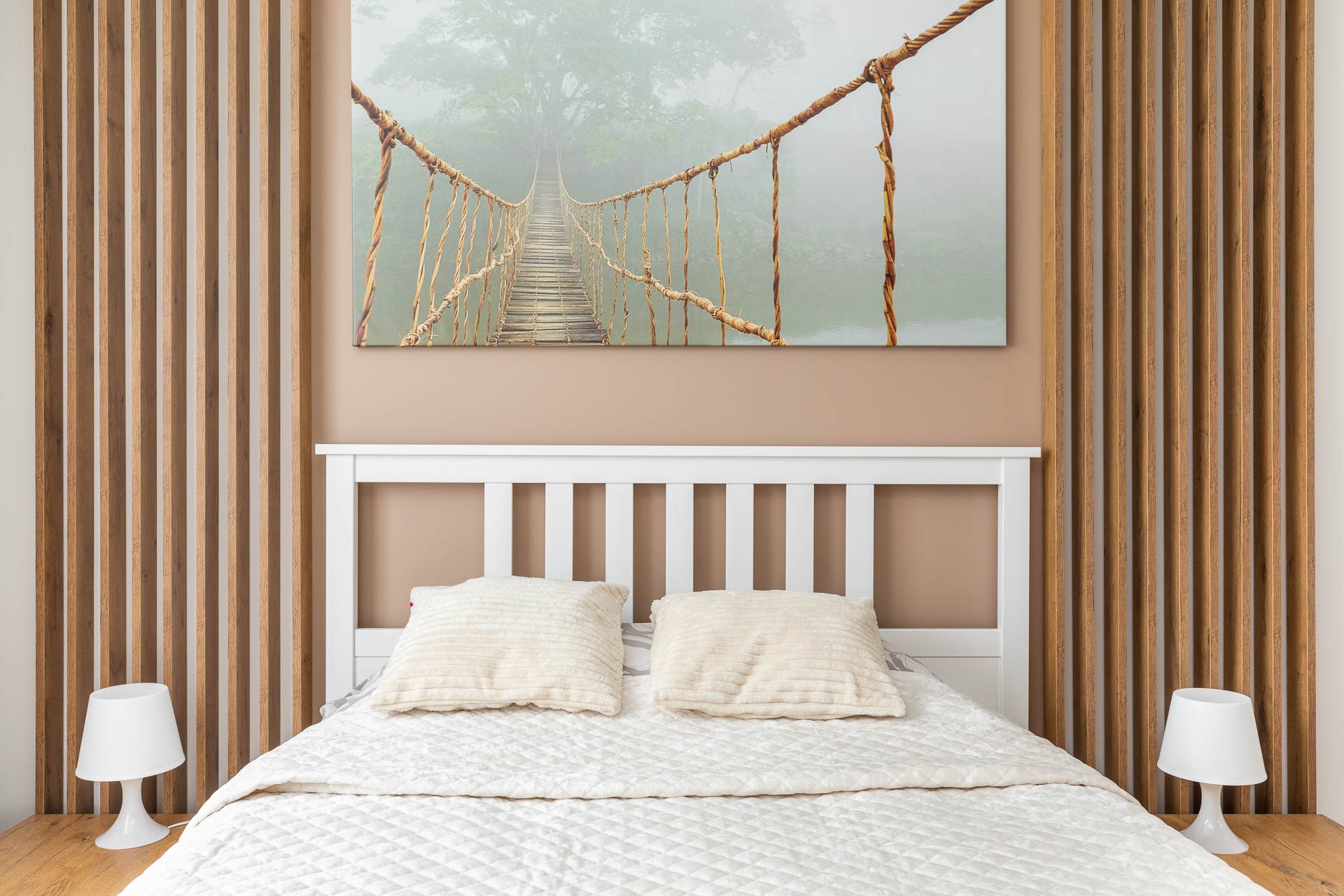 Nowoczesna sypialnia z białym łóżkiem, drewnianą ścianką działową i obrazem z motywem wiszącego mostu w tle.