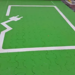 Oznakowane miejsce parkingowe dla samochodów elektrycznych, zielona kostka brukowa z białym symbolem wtyczki i obrysem stanowiska.