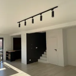 Jasne, przestronne wnętrze po remoncie: białe ściany i sufit, czarna ściana akcentująca, schody, podłoga wyłożona płytkami imitującymi drewno, czarna zabudowa kuchenna w tle i nowoczesne czarne...