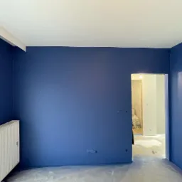 Świeżo pomalowane na granatowo ściany w pokoju, widoczny biały grzejnik i zabezpieczona folią podłoga, przejście do kolejnego pomieszczenia z białymi ścianami.