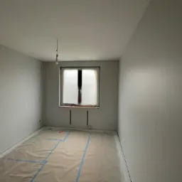 Pokój przygotowany do prac wykończeniowych: ściany po wstępnym wyrównaniu, podłoga zabezpieczona folią z taśmą, okno osłonięte folią, widoczna wisząca żarówka.