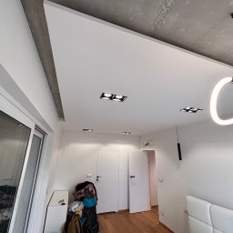 FinitoHome - Nowoczesne wykończenie sufitu w sypialni z oświetleniem punktowym i dekoracyjnym. Widoczna surowa faktura betonu na fragmencie sufitu. Minimalistyczny design wnętrza.