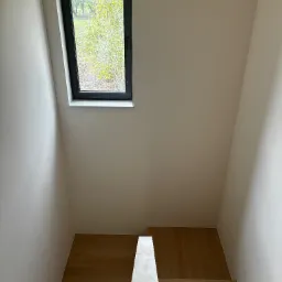 Wnętrze po remoncie: jasne ściany, drewniane schody i czarna rama okienna z widokiem na zieleń. Minimalistyczny design, świeżo pomalowane powierzchnie.