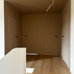 FinitoHome - Korytarz po remoncie: beżowe ściany i sufit, drewniana podłoga. Widoczne drzwi z czarnymi klamkami i wystające kable. Minimalistyczny design.