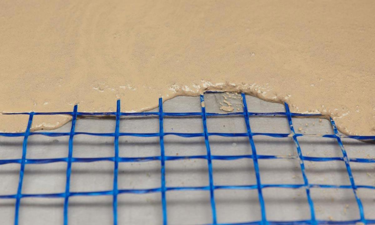 Wylewka samopoziomująca rozprowadzana na siatce pod podłogowe ogrzewanie, widok z bliska na proces zalewania, niebieska siatka, jasnobeżowa masa.