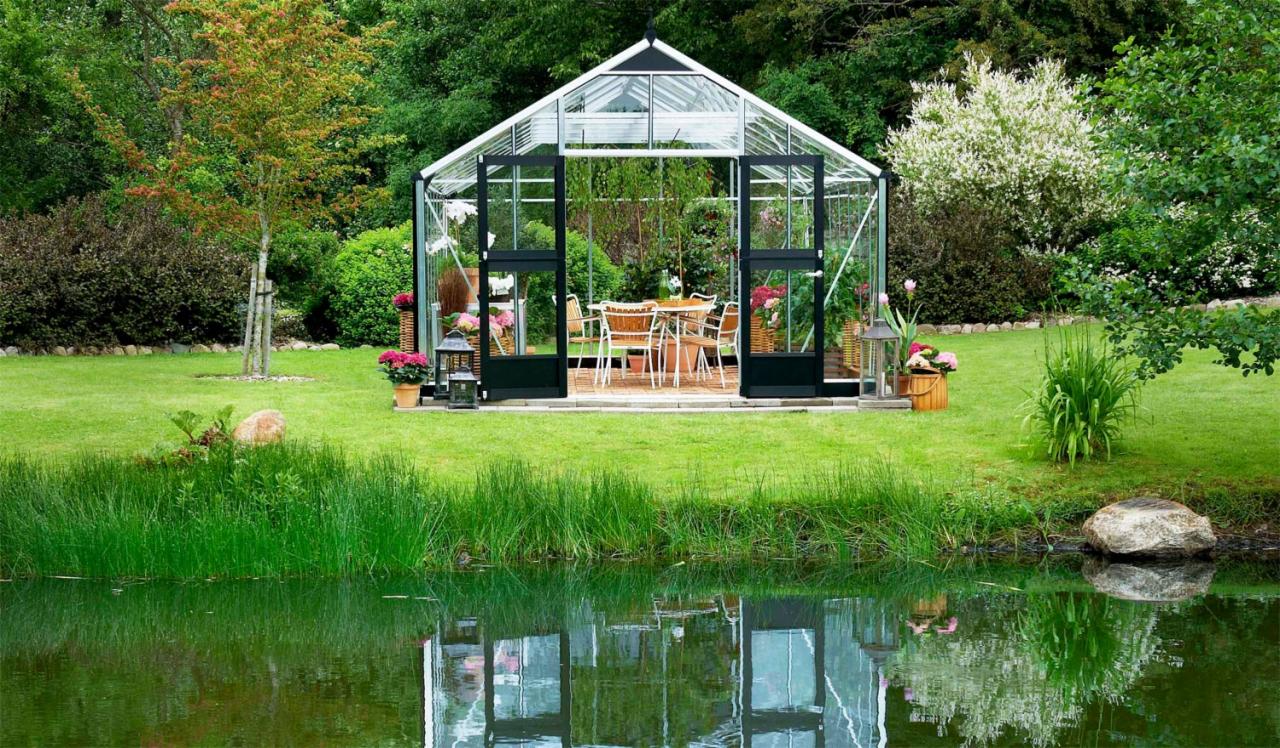 Szklana oranżeria z czarnymi ramami drzwi, otoczona zielonym trawnikiem i stawem, w którym odbija się jej sylwetka.