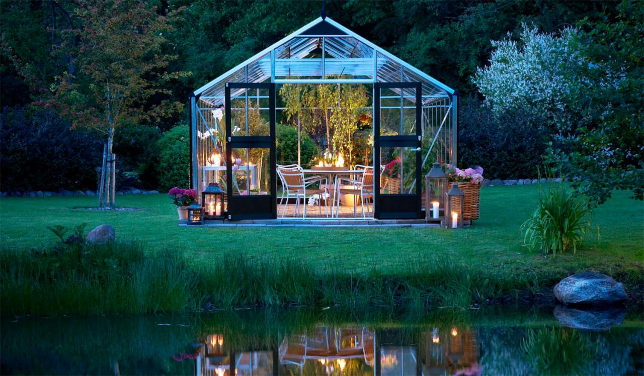 Szklana oranżeria z otwartymi drzwiami, oświetlona lampionami i świecami, z odbiciem w stawie, tworząca romantyczną atmosferę w ogrodzie.