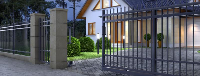 Otwarta, automatyczna brama wjazdowa z metalowymi prętami i kamiennymi słupkami na posesji z kostki brukowej, oświetlona lampami ogrodowymi, z widocznym domem w tle.
