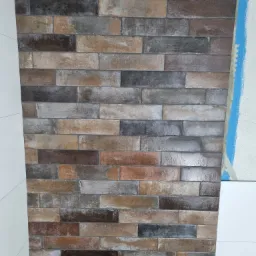 Fragment ściany wyłożonej płytkami imitującymi cegłę w odcieniach brązu i szarości, z widocznym niebieskim oznaczeniem na sąsiedniej ścianie i fragmentem białej płyty po lewej stronie.