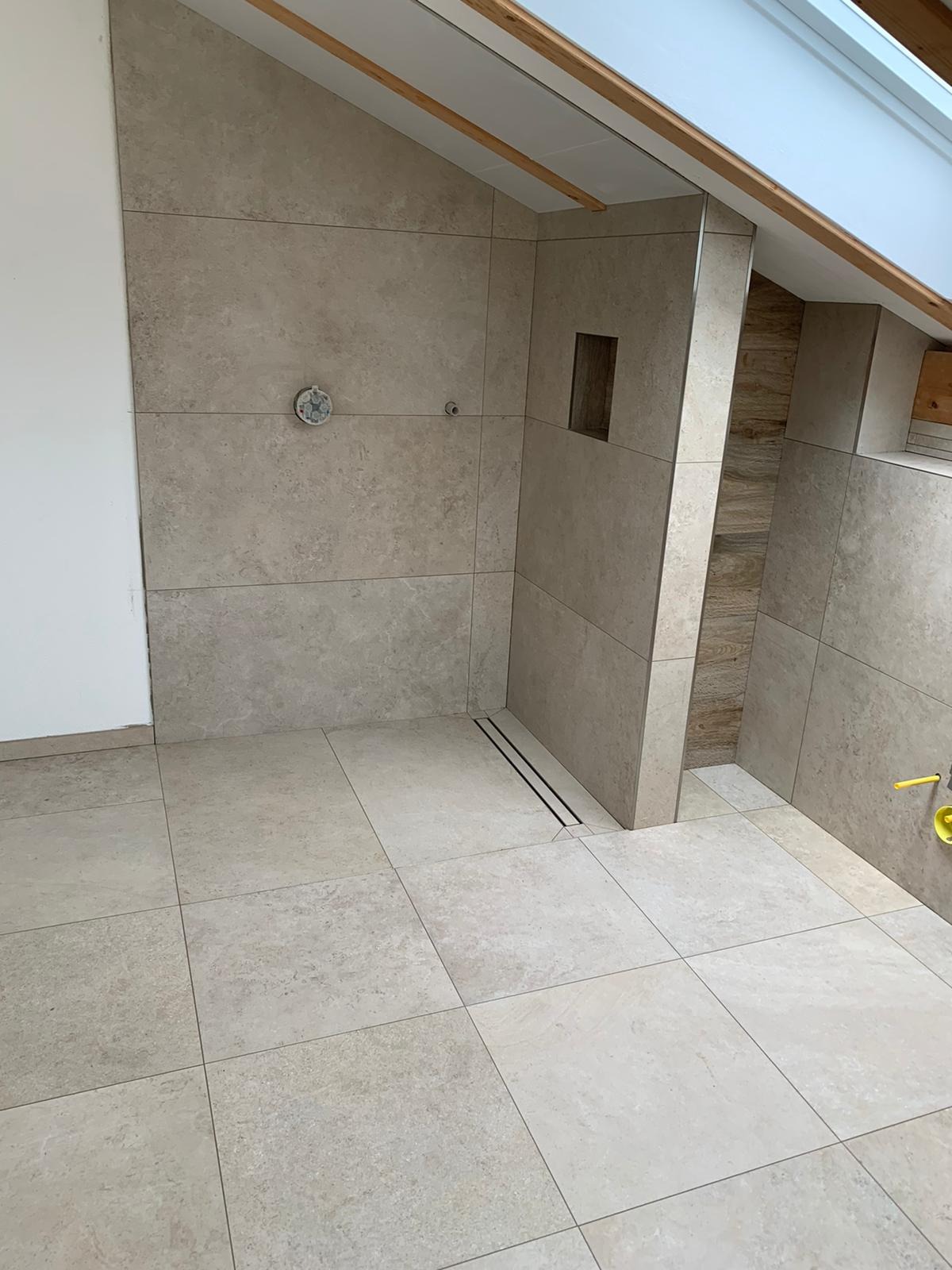 Nowoczesna łazienka na poddaszu z beżowymi, kwadratowymi płytkami ceramicznymi na ścianach i podłodze, z odpływem liniowym w strefie prysznicowej i wnęką w ścianie wykończoną drewnopodobnymi płytkami.