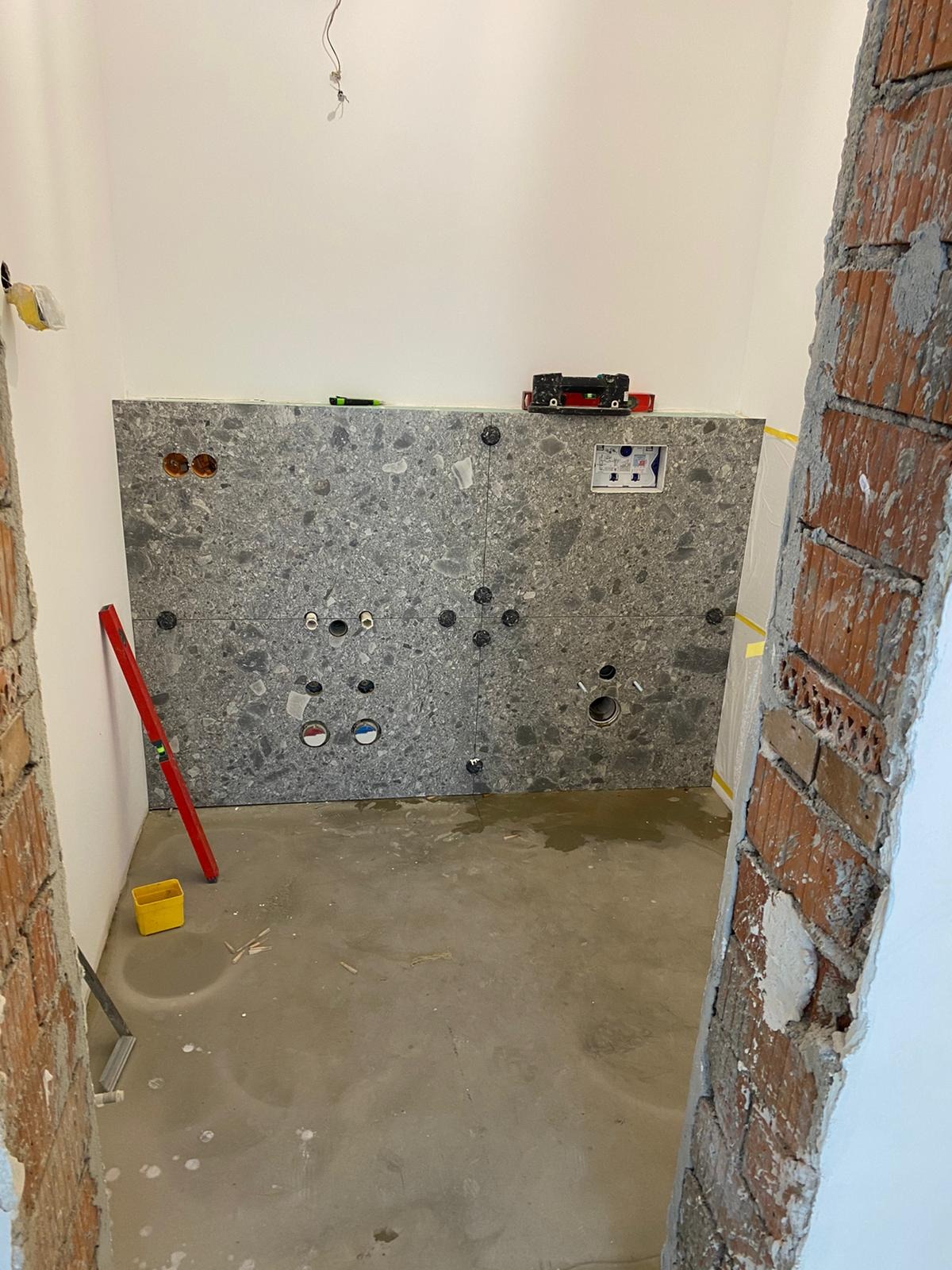 Ściana w trakcie remontu łazienki z zamontowanymi płytami imitującymi lastryko z przygotowanymi otworami na instalacje, widoczny poziomica, żółty pojemnik i surowy stan podłogi.