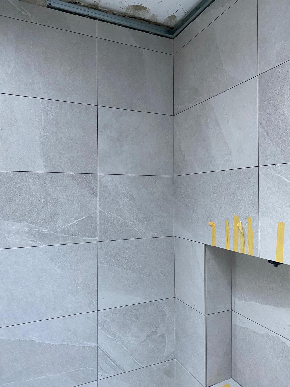Ściana wyłożona szarymi, wielkoformatowymi płytkami imitującymi kamień, z widoczną metalową konstrukcją pod sufitem i taśmą malarską zabezpieczającą krawędzie