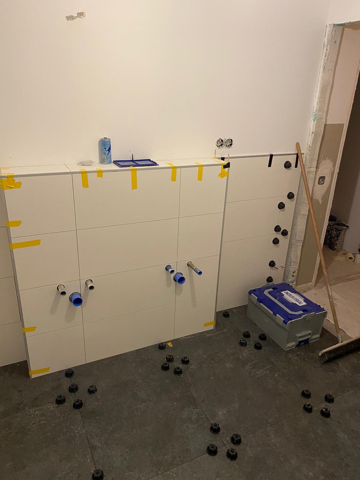 Ściana z białych płytek ceramicznych w trakcie montażu, zabezpieczona żółtą taśmą, widoczne wystające rury i puszki elektryczne, na podłodze szare płytki i czarne podkładki poziomujące, obok...