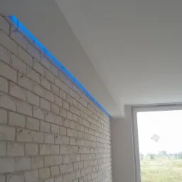 Ściana wykończona cegłą dekoracyjną z podświetleniem LED w kolorze niebieskim, widok z dołu, surowe wnętrze w trakcie wykańczania, okno z widokiem na zewnątrz.