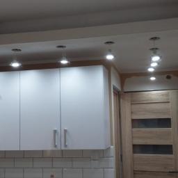 Ad-Instal - Adrian Wiśniewski - Instalacje Elktryczne - Wnętrze kuchni w trakcie remontu: białe szafki wiszące częściowo zakryte folią ochronną, białe płytki na ścianie, rząd punktowych lamp sufitowych, ślady po pracach malarskich na ścianach i sufitach.