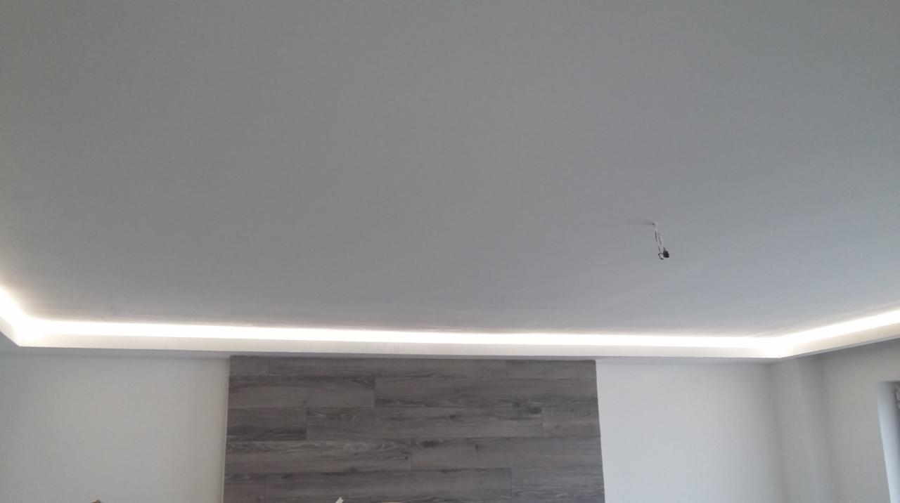 Nowoczesne oświetlenie LED zamontowane wzdłuż krawędzi sufitu w pokoju z dekoracyjną ścianą z paneli imitujących drewno. Widoczne przewody wystające z sufitu w centralnej części.