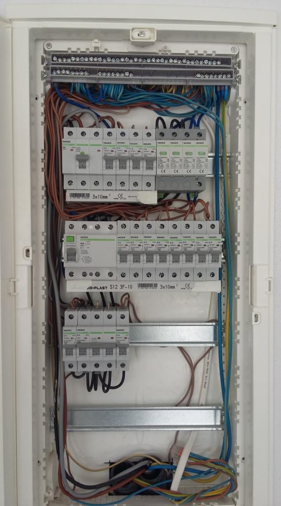 Otwarta skrzynka elektryczna z bezpiecznikami, przewodami i szynami DIN zamontowana na białej ścianie.