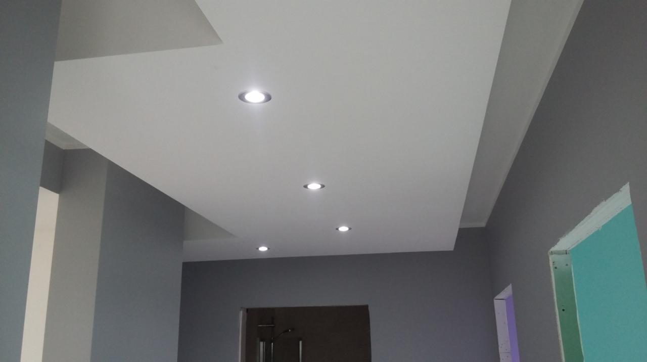 Nowoczesny sufit podwieszany z oświetleniem punktowym LED, szare ściany w tle, widoczne fragmenty konstrukcji sufitu i drzwi.
