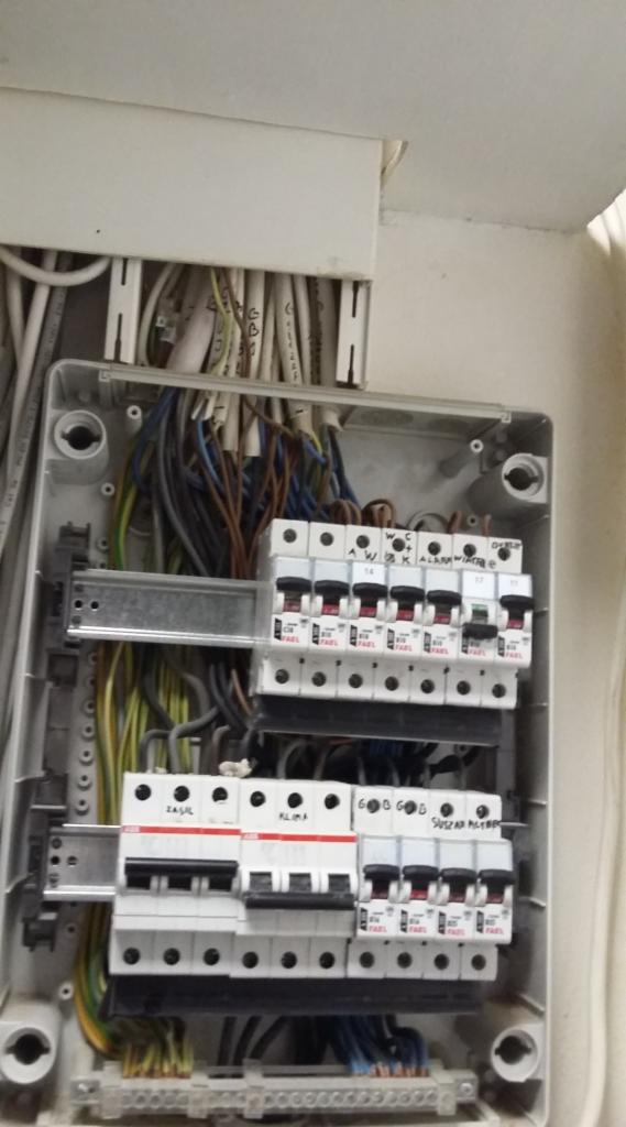 Otwarta skrzynka rozdzielcza z widocznymi bezpiecznikami, przewodami i szynami montażowymi, z ręcznie napisanymi oznaczeniami na niektórych bezpiecznikach.