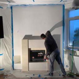 Werbex Sp z o.o. - Pracownik montuje obudowę kominka z jasnego kamienia w zabezpieczonym folią pomieszczeniu, widoczny telewizor na ścianie i okno z widokiem na zewnątrz.