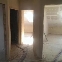 Werbex Sp z o.o. - Wnętrze domu w trakcie wykończenia: widoczne surowe ściany, instalacje elektryczne i przygotowane otwory drzwiowe, z widokiem na poddasze z oknami dachowymi i częściowo zamontowaną płytą karton-gips.