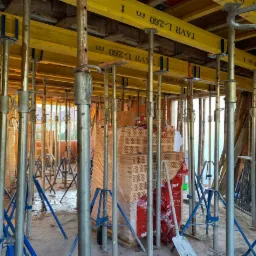 Wnętrze budowy domu w trakcie murowania ścian, widoczne podpory stropowe, ż&oacute;łte dźwigary i paleta z cegłami ceramicznymi w folii.