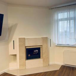 Werbex Sp z o.o. - Salon z jasnymi ścianami, kominkiem obudowanym beżowym kamieniem, telewizorem na ścianie i drewnianą podłogą w odcieniach brązu.