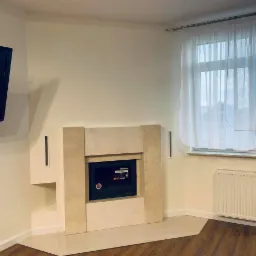 Salon z jasnymi ścianami, kominkiem obudowanym beżowym kamieniem, telewizorem na ścianie i drewnianą podłogą w odcieniach brązu.