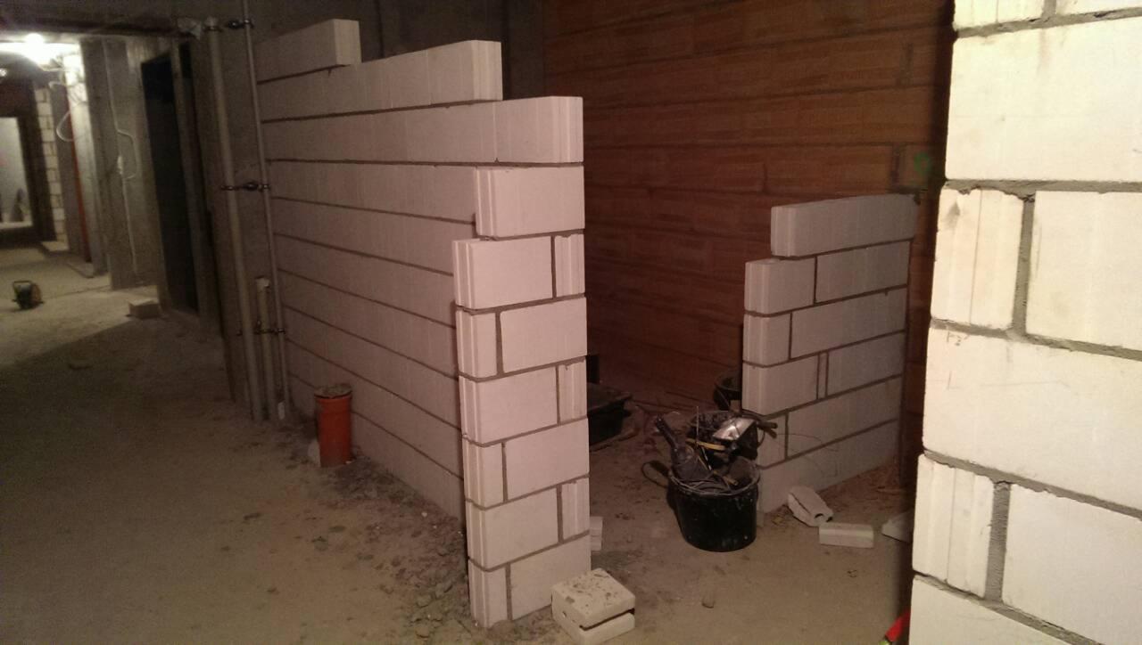 Wznoszenie ścian działowych z białych bloczków w surowym wnętrzu, widoczne narzędzia w wiadrze i fragmenty instalacji.