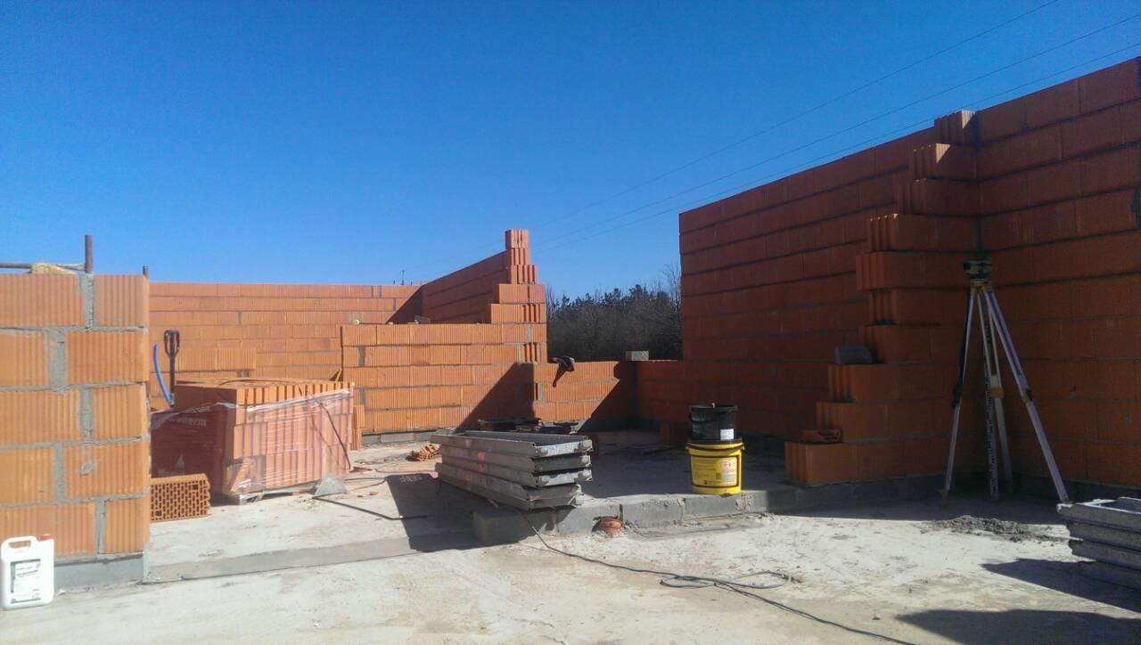 Wznoszone ściany domu z czerwonej cegły ceramicznej, widoczne palety z cegłami, poziomica laserowa na statywie, oraz wiadro z zaprawą murarską na placu budowy w słoneczny dzień.