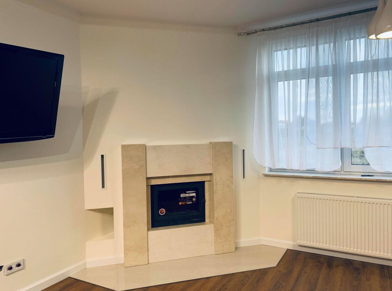 Salon z jasnymi ścianami, kominkiem obudowanym beżowym kamieniem, telewizorem na ścianie i drewnianą podłogą w odcieniach brązu.