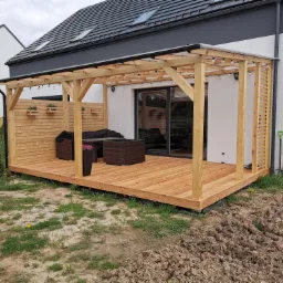 Nowoczesny taras z jasnego drewna z pergolą i oświetleniem, częściowo osłonięty ażurową ścianką, z kompletem mebli wypoczynkowych, widok na dom z szarą dachówką i świeżo przekopaną ziemią w tle.