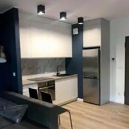 Nowoczesna kuchnia w mieszkaniu w Warszawie, Brylowska. Białe szafki z szarym blatem, marmurowy backsplash, stalowa lodówka i lampy sufitowe punktowe. Widoczny fragment sofy z folią ochronną.