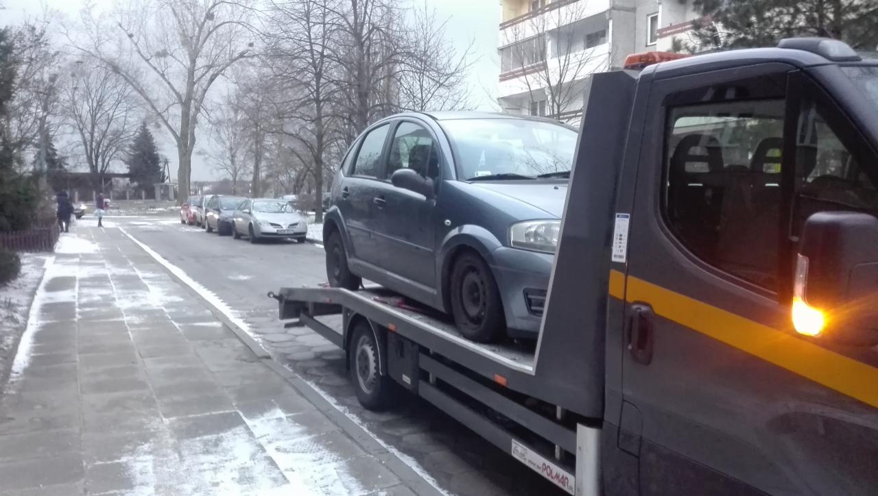 Niebieski Citroen C3 zabezpieczony pasami na platformie lawety na tle zimowego krajobrazu miejskiego z ośnieżonym chodnikiem i rzędem zaparkowanych samochodów.
